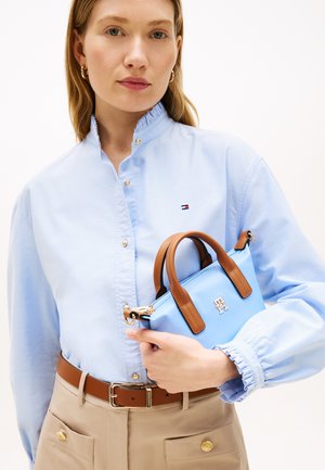 Γυναίκα που φοράει ανοιχτό μπλε μπλούζα και μπεζ παντελόνι, κρατώντας μια ανοιχτό μπλε τσάντα χειρός με καφέ λαβές και χρυσό λογότυπο.