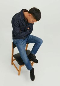 Giacca di denim e jeans blu indossati, seduto su una sedia di legno. Giacca scura con tasche, texture morbida e scarpe in suede nero.