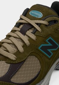Παπούτσι για τρέξιμο New Balance σε ελιά πράσινου χρώματος με μπεζ κορδόνια, διχτυωτά πάνελ και μαύρο λογότυπο "N" με μπλε περίγραμμα στο πλάι.