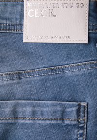 Tissu en denim bleu clair avec une texture subtile, doté d'une étiquette en cuir blanc embossée portant le nom de la marque "Cecil" et des coordonnées géographiques.