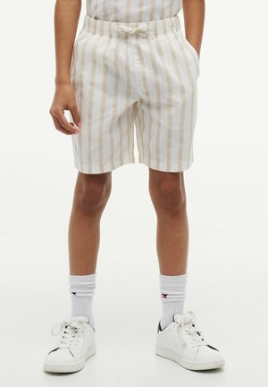 Niño vestido con pantalones cortos de rayas verticales beige y blancas con cintura elástica, calcetas blancas hasta la pantorrilla y zapatillas blancas, de pie con las manos en los bolsillos.