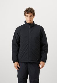 Young man with curly hair wearing a black zip-up puffer jacket and black pants, standing against a plain white background.