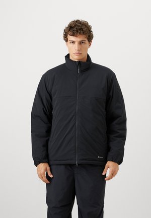 Young man with curly hair wearing a black zip-up puffer jacket and black pants, standing against a plain white background.