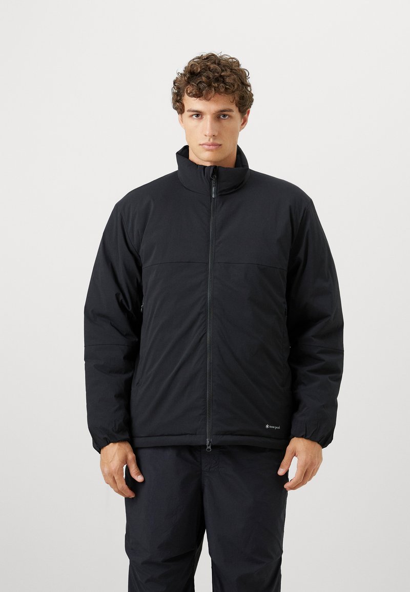 Young man with curly hair wearing a black zip-up puffer jacket and black pants, standing against a plain white background.