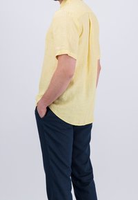Chemise en lin jaune clair à manches courtes, col avec un ourlet arrondi et une poche poitrine unique, associée à un pantalon bleu marine foncé.