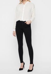 Camisa blanca de manga larga con cierre de botón, combinada con jeans negros ajustados y zapatos de tacón alto negros. Tela suave y corte entallado.