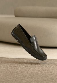Mocassin en cuir noir avec semelle en caoutchouc texturée flottant au-dessus d'un tapis beige et d'un mobilier rembourré assorti en arrière-plan.
