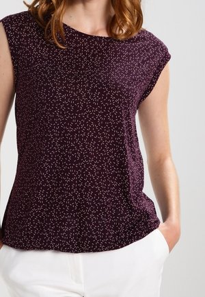 Kobieta ubrana w bezrękawnik w ciemnym fioletowym kolorze z małym wzorem drobnych jasnoróżowych kresek, w połączeniu z białymi spodniami, z ręką w kieszeni.