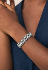 Sølvfarvet metalarmbånd med sammenkædede rektangulære led, glat overflade og fleksibel design, båret om håndleddet.