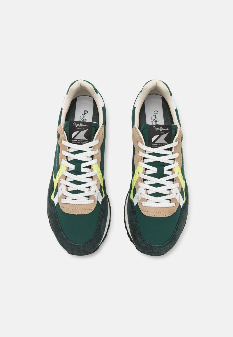 Zapatillas deportivas verdes y beige con cordones blancos, de materiales de ante y textil, que presentan un acento amarillo y suela de goma negra. Diseño atlético clásico.