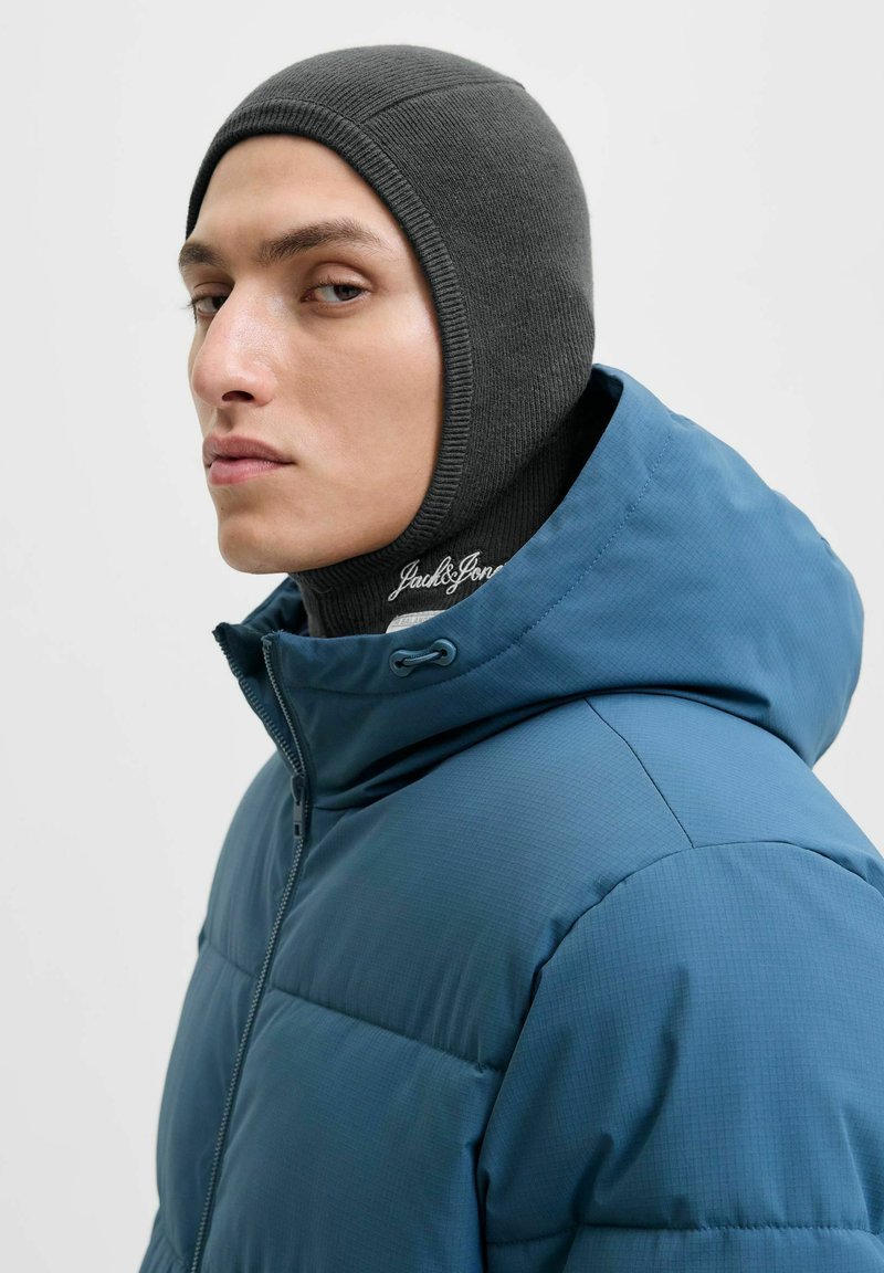 Jeune homme portant une cagoule grise et une doudoune à capuche bleue, regardant sur le côté avec une expression neutre.