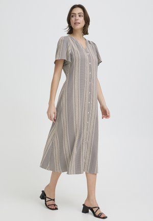 Nő bézs és fekete mintás midi ruhában, fekete magassarkú szandállal, egyszínű világos háttér előtt sétál.