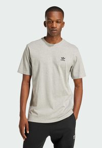 Siva bombažna majica s kratkimi rokavi, okroglo izrezano ovratnikom in črnim logotipom Adidas na prsih. Obuta z črnimi trenirkami.