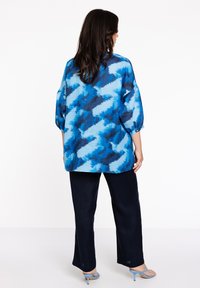 Helles Oberteil mit blauem Muster und Ballonärmel, kombiniert mit dunkelblauen Hosen und hellblauen Riemchensandalen. Glattes Material mit abstrahierten Designelementen.