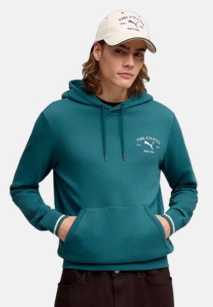 Jonge man met een turquoise Puma hoodie en een beige Puma pet, handen in de zak van de hoodie, kijkt lichtjes opzij met een neutrale uitdrukking.