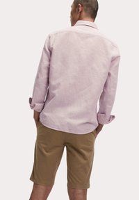 Camisa de manga larga rosa claro con cuello tipo abotonado y puños enrollados, combinada con pantalones cortos beiges. La tela parece texturizada y casual.