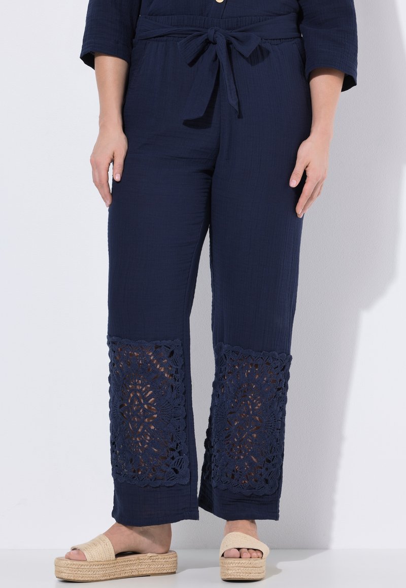 Pantalones azul marino con cintura anudada y piernas anchas que presentan paneles de encaje intrincados sobre sandalias de plataforma beige.