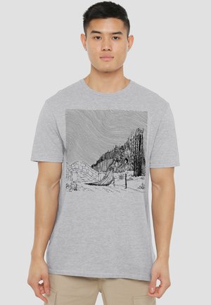 Man wearing light gray T-shirt with detailed black line art of a figure in a flowing dress walking on a path near trees and hills.