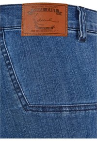 Sinised denim teksad, millel on pruun nahklabel, millel on reljeefne tekst. Materjalil on õrn tuhmus ja õmmeldud detailid taskutel.