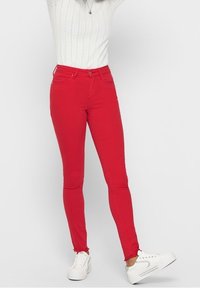 Vaqueros skinny rojos con una textura suave, que cuentan con bolsillos frontales, presillas para cinturón y un cierre de botón. Combinados con una blusa blanca y zapatillas blancas.