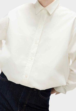 Osoba ubrana w białą koszulę na guziki wpuszczoną w ciemnoniebieskie dżinsy, z rękami złożonymi za plecami.