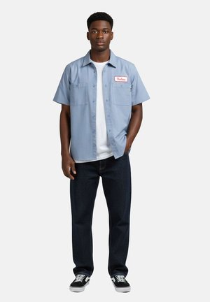 Jeune homme portant une chemise à manches courtes bleu clair boutonnée par-dessus un t-shirt blanc, un jean foncé et des baskets noires à lacets blancs.