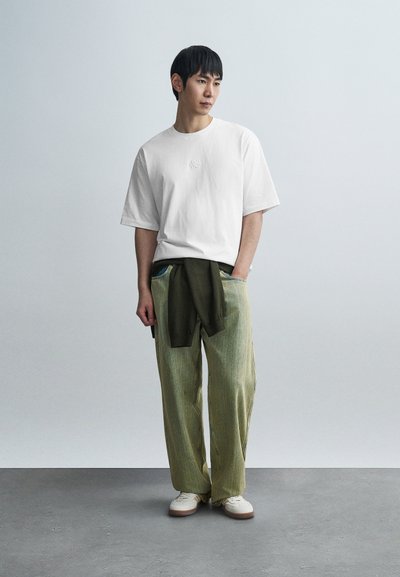 Λευκό υπερμεγέθες μπλουζάκι με μικρό λογότυπο, σε συνδυασμό με φαρδιά παντελόνια με ανοιχτό πράσινο ρίγες και λευκά παπούτσια με καφέ σόλες.