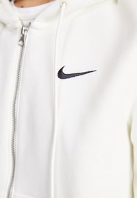 Vit zip-up huvtröja i mjukt material, med en svart Nike swoosh-logotyp på bröstet. Inkluderar huva med dragsko och silverfärgad dragkedja.