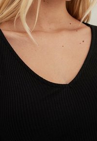 Černá žebrovaná tkanina má výstřih do V, s mírně texturovanými detaily a těsným střihem. Ton pleti viditelný na výstřihu a ramenou.