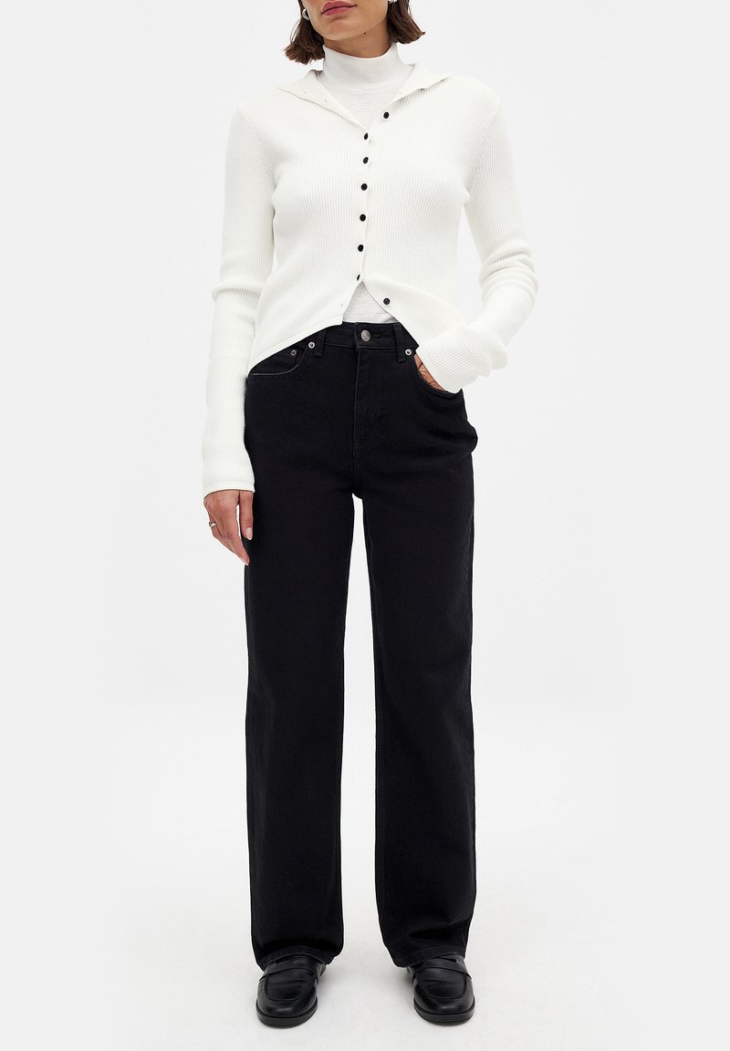 Cardigan blanco de punto acanalado con capucha, sobre un cuello de cisne blanco, combinado con jeans de pierna ancha negros de cintura alta y zapatos negros.