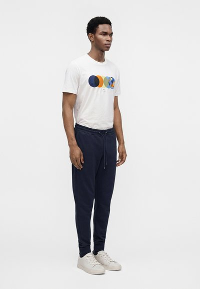 Νέος άνδρας όρθιος, φοράει λευκό t-shirt με πολύχρωμα κυκλικά γραφικά, σκούρο μπλε παντελόνι jogger και λευκά αθλητικά παπούτσια μπροστά σε ένα απλό φόντο.