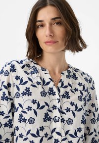 Hvid bluse med marineblå blomsterbroderi, rund halsudskæring og knaplukning. Blødt, letvægtsstof med synlige sy-mønstre.