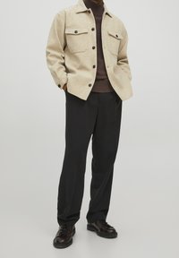Beige wool blend shirt jacket with two chest pockets, brown ribbed sweater underneath, black trousers, and shiny black lace-up boots.