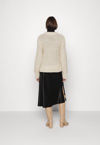 Flauschiger cremefarbener Strickpullover mit gerippten Bündchen, kombiniert mit einem schwarzen Satin-Nachtkleid mit seitlichem Schlitz und beigefarbenen Kniehohen Stiefeln mit Absatz.