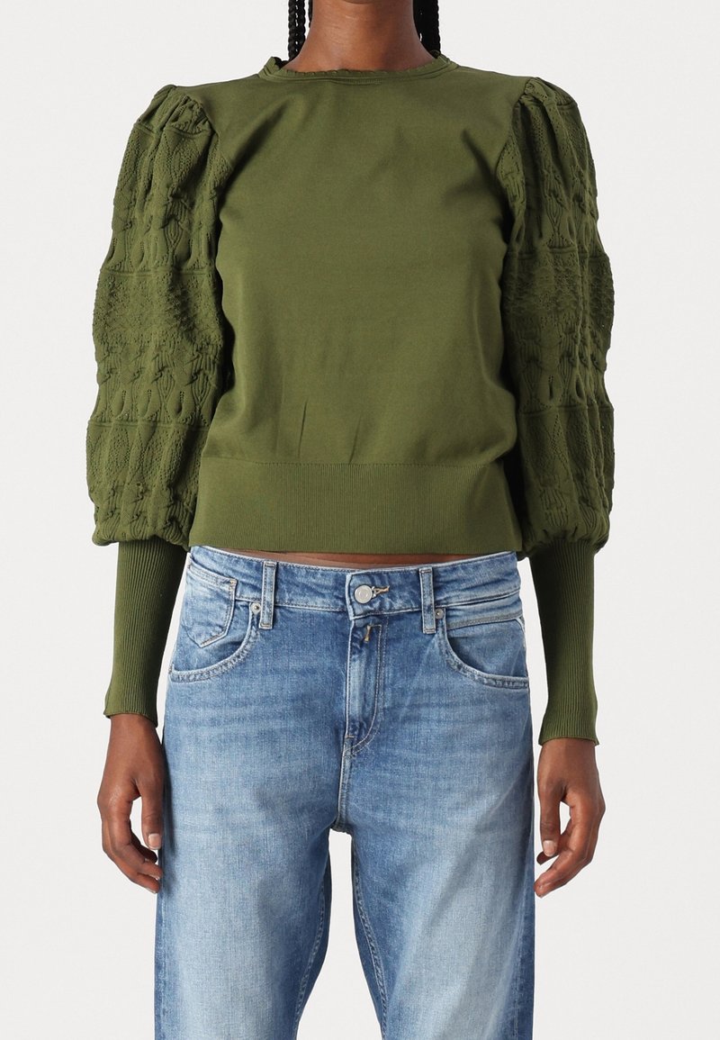 Personne portant un pull vert olive avec des manches bouffantes texturées et un jean bleu, debout devant un fond clair uni.