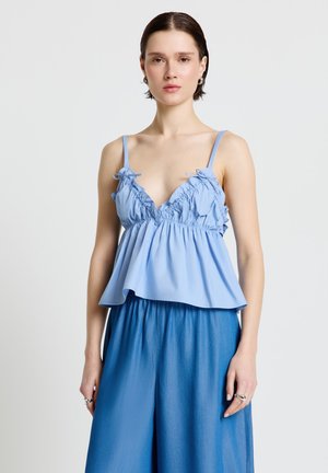 Femme portant un haut sans manches bleu clair à volants et un pantalon large bleu, debout devant un fond blanc avec une expression neutre.