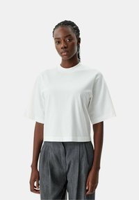 Witte cropped t-shirt met korte mouwen, met een subtiele "PINKO" logo in bijpassende draad. Zachte stof en een ontspannen pasvorm. Gecombineerd met grijze broeken.