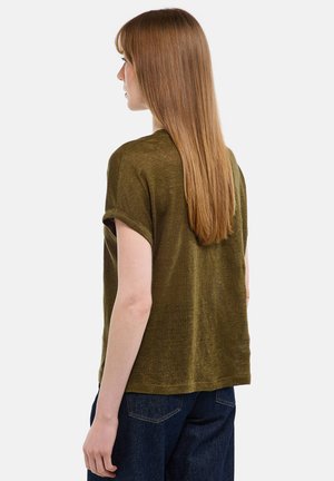 Femme aux cheveux longs et lisses portant un haut à manches courtes vert olive et un jean bleu foncé, vue de dos sur un fond blanc.