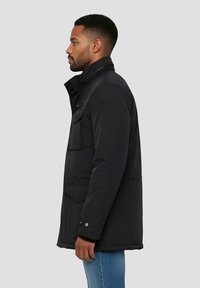 Giacca nera isolata con colletto alto, tasche frontali e chiusura con bottoni. La texture appare liscia, con un design elegante.