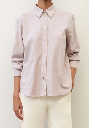 Femme portant une chemise beige clair à manches longues avec boutons et col, associée à un pantalon blanc cassé, debout devant un fond uni.
