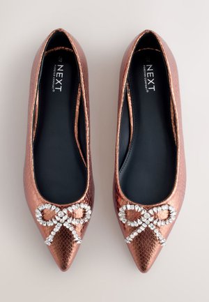 Zapatos planos puntiagudos en oro rosa con un patrón texturizado de piel de reptil, con un lazo adornado con cristales en la punta. Forro interior negro.