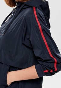 Navy windbreaker with red side stripes, hood, and elastic cuffs. Smooth, lightweight material; features a front zipper pocket and adjustable drawstrings.