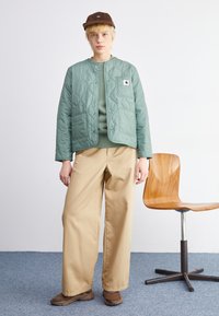 Mintgroene gewatteerde jas met drukknoopjes en twee voorzakken, gecombineerd met een groene top, beige wijde broek en bruine schoenen.