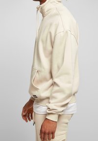Uomo che indossa un felpa a collo alto color crema e pantaloni della tuta abbinati, con le mani rilassate lungo i fianchi, inquadrato a partire dal mento e dalle ginocchia.
