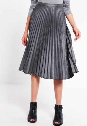 Femme portant une jupe midi plissée gris métallique associée à des bottines noires à bouts ouverts, debout devant un fond uni.