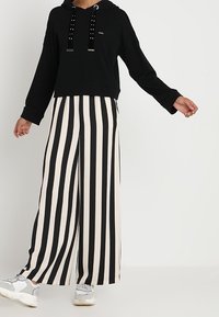 Felpa nera con lacci borchiati e design accorciato, abbinata a pantaloni a gamba larga con righe verticali nere e crema.