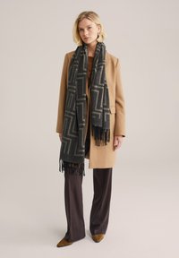 Pelēks un melns rakstains šalle ar bārkstīm, uzvilkta virs brūnganas mēteļa. Ievērojams ģeometrisks dizains. Modelis apvienots ar tumši brūnām biksēm un norādītiem apaviem.