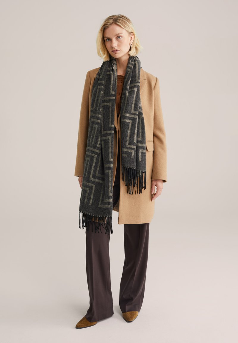 Pelēks un melns rakstains šalle ar bārkstīm, uzvilkta virs brūnganas mēteļa. Ievērojams ģeometrisks dizains. Modelis apvienots ar tumši brūnām biksēm un norādītiem apaviem.