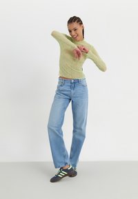 Lys grønn, gjennomsiktig langermet topp med bølgende tekstur, kombinert med mellomhøye, lyseblå jeans og marineblå sko med grønne striper.