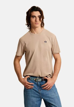 Jeune homme aux cheveux mi-longs portant un t-shirt beige et un jean bleu avec une ceinture à motifs, mains posées sur les hanches.